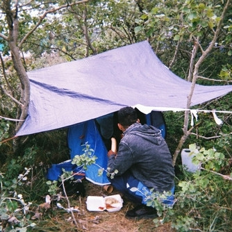 Young person sitting under tarpaulin draped between trees eating food